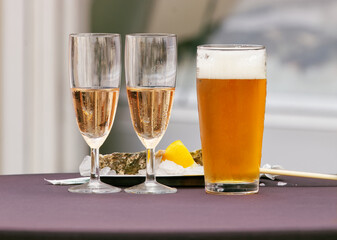 Close-up of two champagne glasses and a pint of beer placed on a table with oysters and lemon wedge, refreshing drinks at outdoor event.