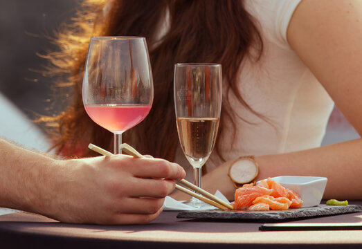 Close-up of couple enjoying sushi with chopsticks and glasses of wine on outdoor table, romantic dining moment.