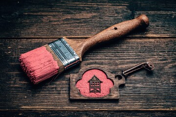 A paintbrush with pink paint, resting on a weathered wooden surface, next to a small wooden house charm