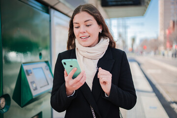 Cheerful female actively engaged in using her smartphone to browse applications while waiting at a transportation station, showcasing her excitement and connection to technology in a busy setting