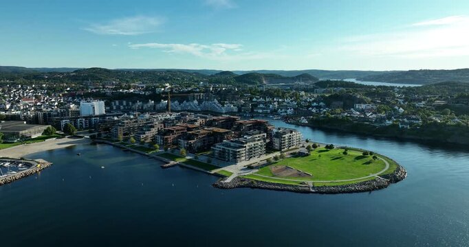 Bright sunny day showcases Kristiansand in Norway with modern architecture, a serene marina, and lush green areas along the waterfront. Scenic hills rise in the background.
