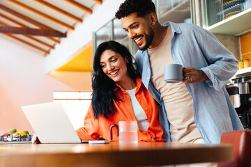 Smiling couple enjoying morning coffee while browsing on a laptop in kitchen