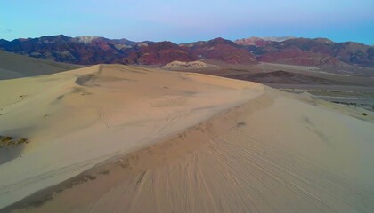 Naklejka premium A vast expanse of beige sand dunes stretches towards a backdrop of colorful mountains under a serene, pale blue sky.