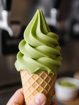 A realistic close-up of vibrant green matcha soft-serve ice cream being swirled into a wafer cone held by a hand