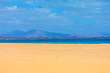 Wide sandy beach in Fuerteventura stretches toward turquoise waters and distant volcanic mountains. Bright sky and open space create a tranquil Canary Islands landscape