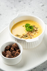 Creamy pumpkin soup in a white bowl, garnished with seeds and parsley, served with croutons on a white plate over a speckled gray background