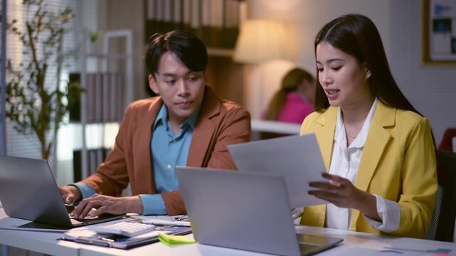 Two people are sitting at a desk with laptops open, one of them is a woman in a yellow jacket. They are looking at a piece of paper and discussing something