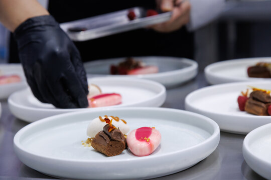 plated desserts in professional restaurant kitchen. chef arranges chocolate cake, mousse, and artistic garnishes on plates. fine dining presentation, culinary art and gourmet food preparation.