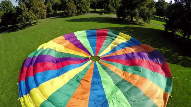 Aerial view of a vibrant, multicolored parachute lying on a lush green field with trees in the background.