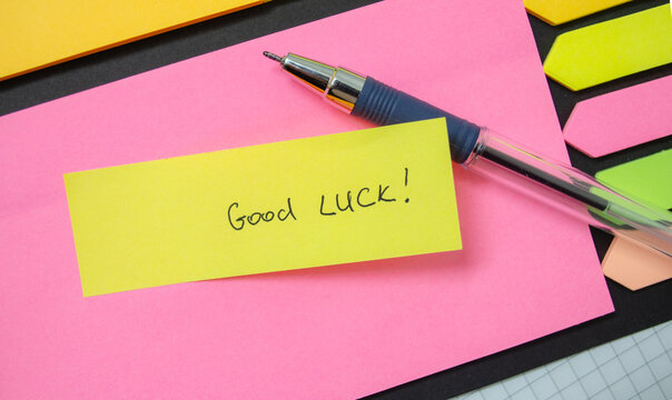 Bright sticky notes in pink and yellow lay on a table alongside a pen, featuring a handwritten message wishing good luck, ideal for motivating study or work sessions. Copy space.