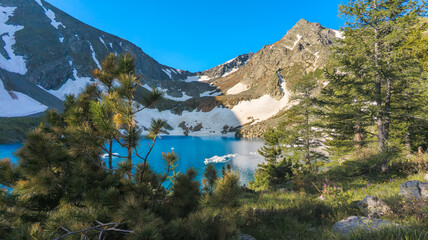 A serene alpine lake in a mountainous setting. Snow-capped peaks surround the deep blue water. Pine trees and clear blue sky create a peaceful, awe-inspiring scene.