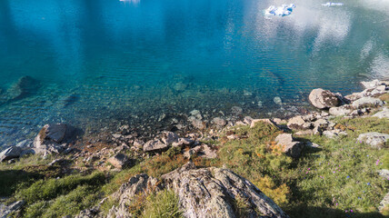 An image of a serene turquoise lake with visible rocks at the bottom, surrounded by rocks, wild grass, and boats, set against a clear blue sky with mountains in the background