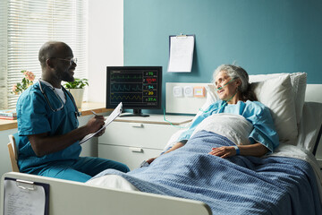 Caucasian senior woman lying in hospital bed interacting with Black male doctor wearing stethoscope, doctor holding clipboard and writing notes, medical monitor displaying vital signs