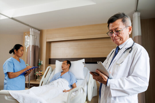 asian senior male professional doctor using tablet for digital health record and nurse writes notes beside patient. hospital room equipped medical instruments and IV drip. healthcare and treatment. - Powered by Adobe