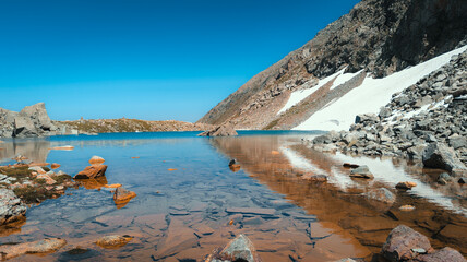 Serene mountain lake reflects turquoise waters under a clear blue sky, surrounded by rocks and snowy mountains, creating a tranquil setting.