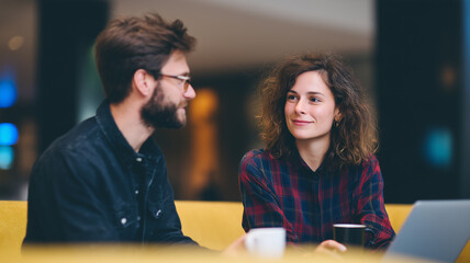 Young colleagues discussing ideas on yellow sofa in modern hotel lobby, smiling and enjoying coffee together