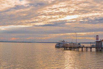 Obraz premium Lakeside sunset bar on a pier with a panoramic view of Podersdorf Lighthouse at Lake Neusiedl, Austria, during golden evening light.