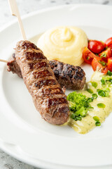 Grilled meat skewers with mashed potatoes, cherry tomatoes, and creamy herb sauce on a white plate over a gray speckled surface
