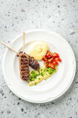 Fototapeta na wymiar Grilled meat skewers with mashed potatoes, cherry tomatoes, and creamy herb sauce on a white plate over a gray speckled surface