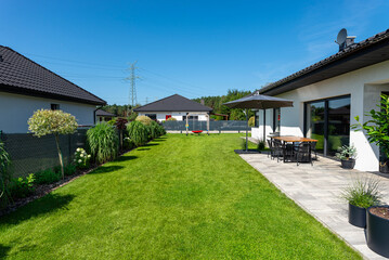 A beautiful garden with green grass, a patio table and umbrella visible, a house in beautiful summer weather, a very wide shot.