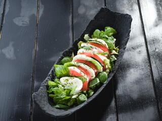 Delicious Caprese salad, featuring sliced tomatoes and mozzarella, is garnished with fresh basil leaves and served in a decorative black platter on a dark wood table