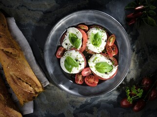 A rustic metal plate holds Caprese salad with fresh basil. Next to it, a linen cloth supports crusty bread. Red chili peppers add a spicy touch to this colorful and tempting appetizer