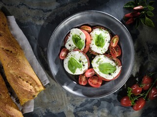 Caprese salad is displayed on a pewter plate, featuring ripe tomatoes, creamy mozzarella, fresh basil, and cracked pepper, complemented by rustic bread and small tomatoes on the vine