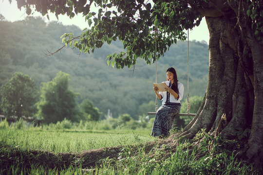 Beautiful Asian woman in traditional dress enjoys reading a book on a swing while enjoying nature and fresh air in a rice field in Isan, Thailand.