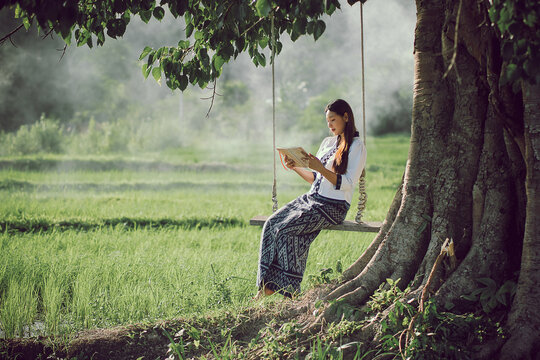 Beautiful Asian woman in traditional dress enjoys reading a book on a swing while enjoying nature and fresh air in a rice field in Isan, Thailand.