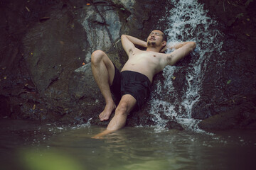 A Young Asian man in lotus yoga pose meditating on a peaceful natural waterfall. Beautiful natural sculpture of art and religion.