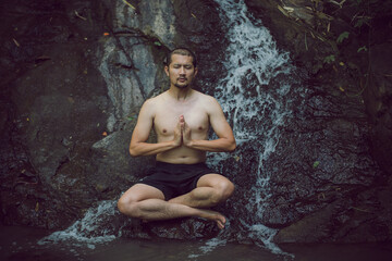 A Young Asian man in lotus yoga pose meditating on a peaceful natural waterfall. Beautiful natural sculpture of art and religion.