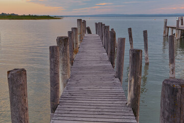 Fototapeta premium Wooden pier leading over a serene lake with soft pastel sunset sky, water reflecting warm orange and pink tones, creating an atmospheric and mysterious evening