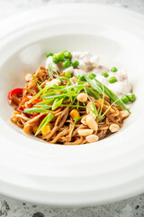 Asian noodle salad with vegetables, peanuts, and green onions, served with creamy white sauce and green peas on a white plate over gray stone background.