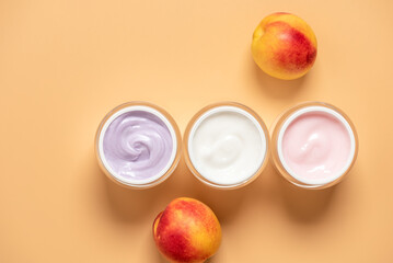 Set of cosmetic creams of different colors in open jars with fresh peaches on yellow pastel background, top view, rejuvenating, moisturizing, organic creams