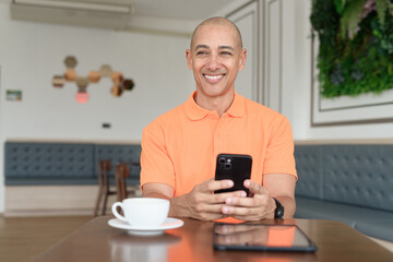 Bald Hispanic businessman having coffee break in cafe and using phone while thinking