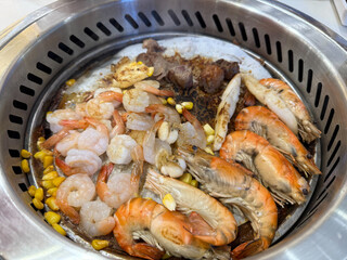 Various seafood and meats, including shrimp, are grilled on a Korean-style barbecue pan with corn and garlic, arranged appetizingly in a round stainless steel grill