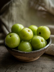 A rustic ceramic bowl overflowing with vibrant green indian gooseberries also known as amla rests on a weathered wooden surface