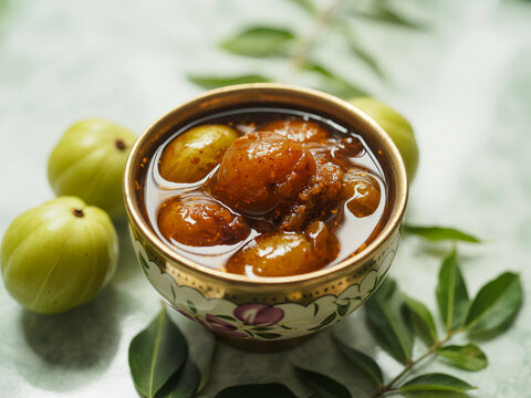 A close up of indian gooseberry murabba in a decorative bowl surrounded by fresh gooseberries and green leaves