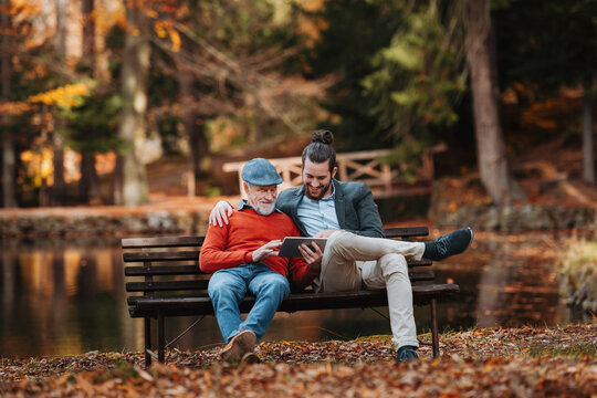 Senior father and his son sitting on bench by lake in nature, looking at tablet.