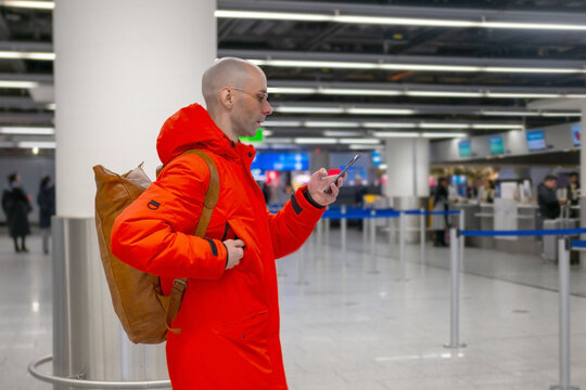 Man in orange jacket, Modern Solo traveler checking flight status on phone in terminal, Passenger with backpack in Airport Germany using smartphone near gate, Frankfurt - February 10, 2025