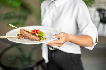 Person holding plate with grilled sausages on skewers, served with cherry tomatoes, broccoli, and cucumbers. Outdoor setting, casual dining presentation