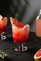 Cocktail or mocktail with grapefruit tonic and red grenadine syrup in a stemmed glass garnished with rosemary. Dark wooden table, palm tree leaves in the background. Two glasses of ice citrus drink