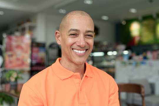 Bald Hispanic man happy smiling face inside cafe restaurant wearing orange polo shirt