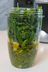 A clear blender cup is filled with layers of raw ingredients, including pistachios, parmesan cheese, and fresh basil leaves, ready for blending.