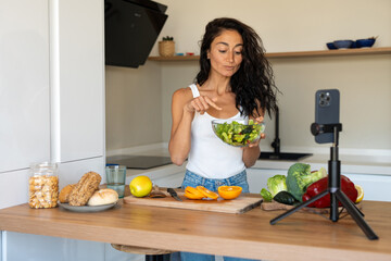 Brunette woman in white tank top pointing at a fresh green salad bowl while filming a healthy cooking video in a modern kitchen with vegetables on the table.