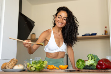 Fit and smiling woman in a white sports top tasting fresh vegetable salad with chopsticks in a modern kitchen, surrounded by healthy food ingredients and vegetables.