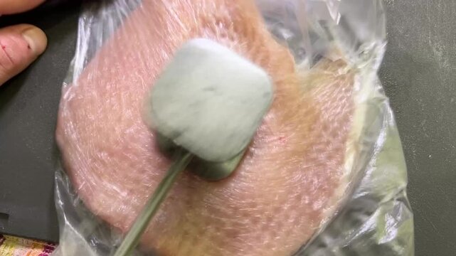 Flattened raw chicken breast under plastic wrap being tenderized with a metal mallet