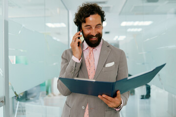 Happy businessman reading notes and talking on mobile phone while walking in modern office