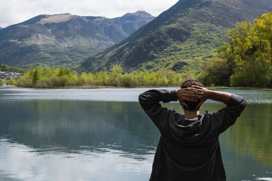 Man Contemplating Nature's Serenity - A lone man sits by a pristine lake, captivated by the breathtaking beauty of surrounding mountains and their reflections in the tranquil water.