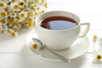 Aromatic tea in cup, chamomile flowers and spoon on white wooden table, closeup